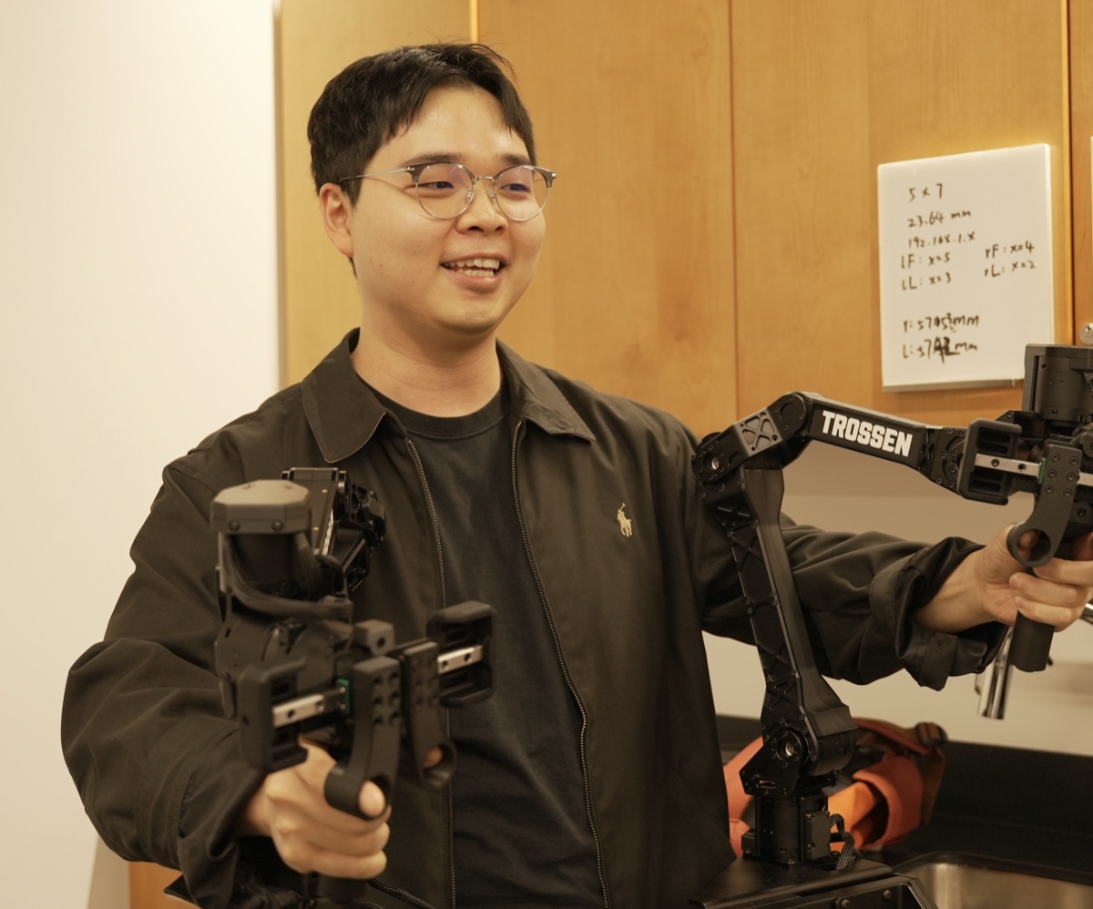 Seungjae Lee, holding teleoperation grippers beside a robot arm